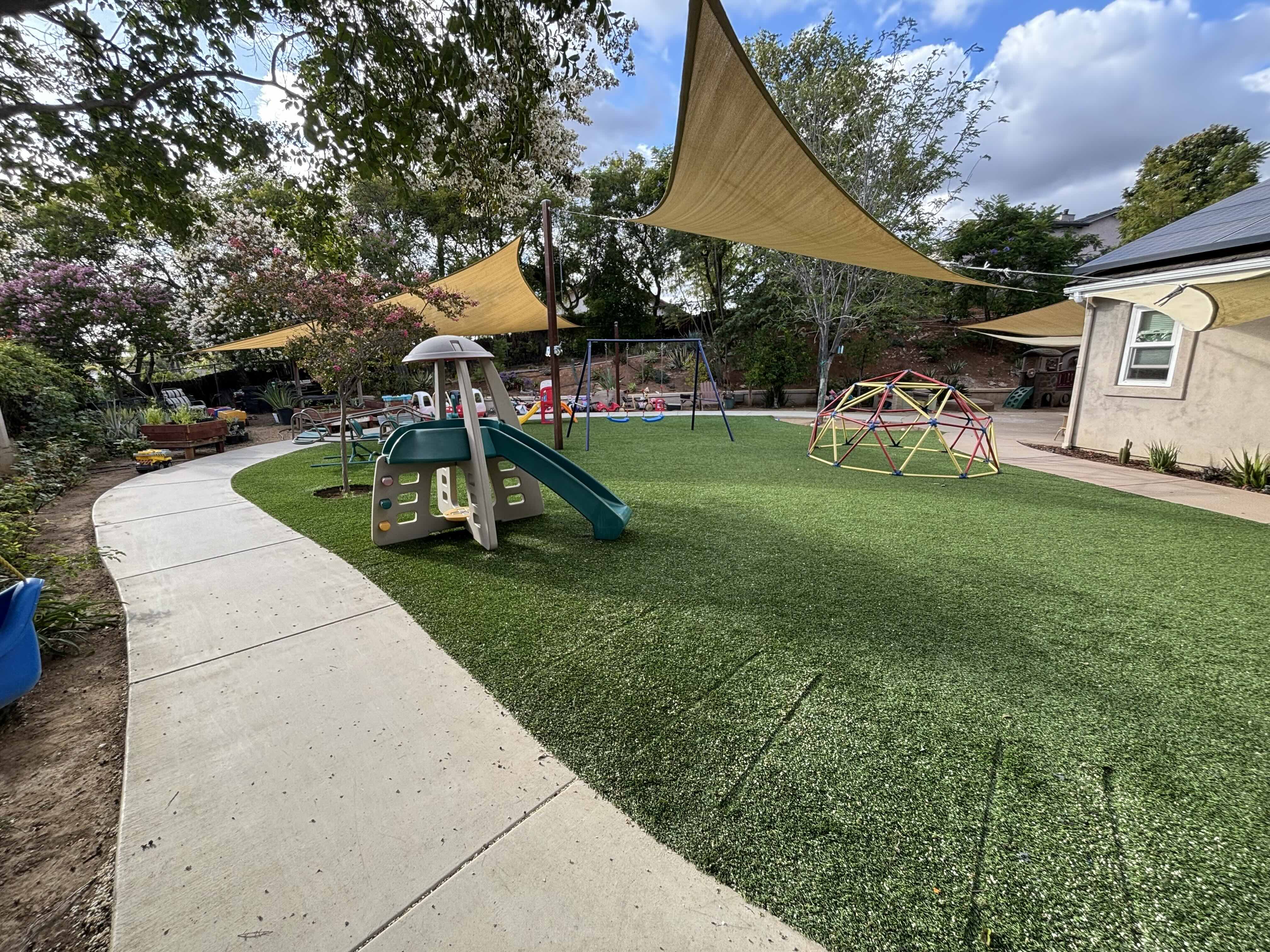 The backyard playspace at Ms. Rena's Preschool in Escondido, California — featuring a concrete bike path, jungle gym, climbing dome, and shade sails over artificial turf